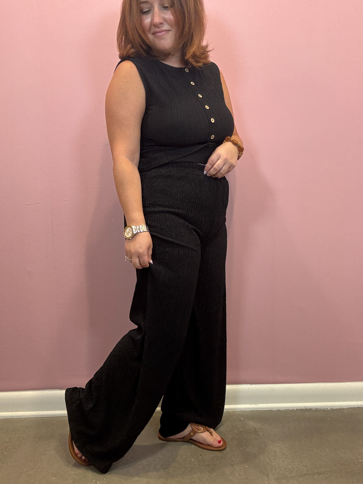 A woman modeling a black sleeveless jumpsuit with gold buttons down the front, paired with brown leather sandals, standing against a pink wall.