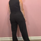 A woman wearing a black sleeveless ribbed jumpsuit, viewed from the back, standing against a pink wall.