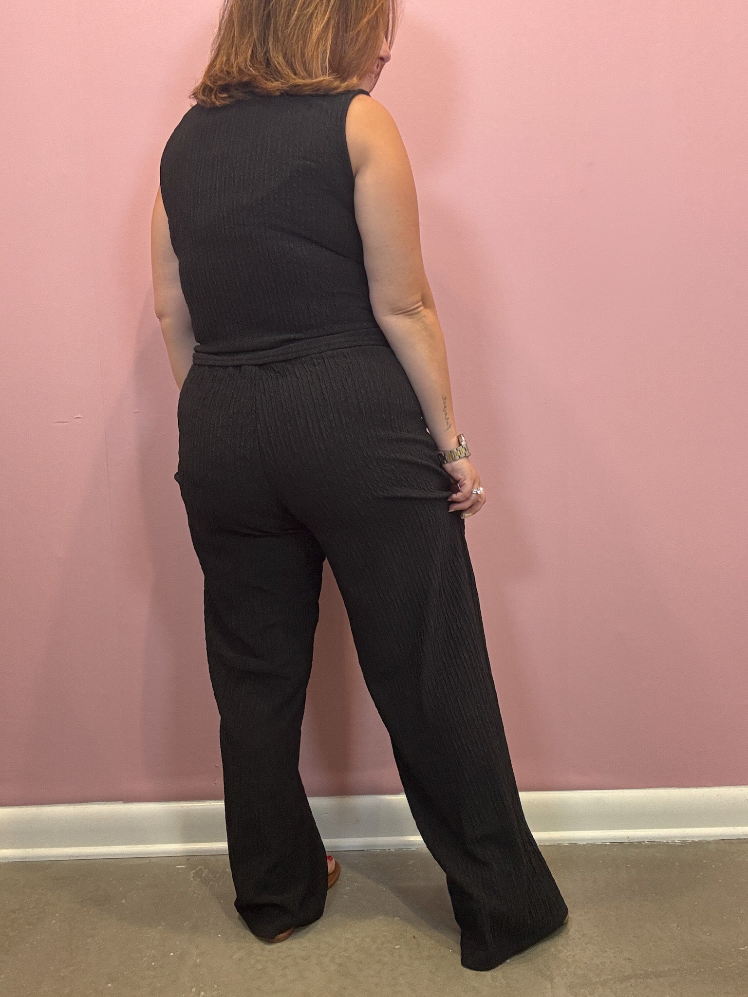 A woman wearing a black sleeveless ribbed jumpsuit, viewed from the back, standing against a pink wall.