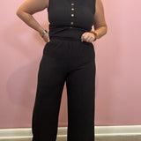 A woman modeling a black ribbed sleeveless top and matching wide-leg pants set against a pink wall.