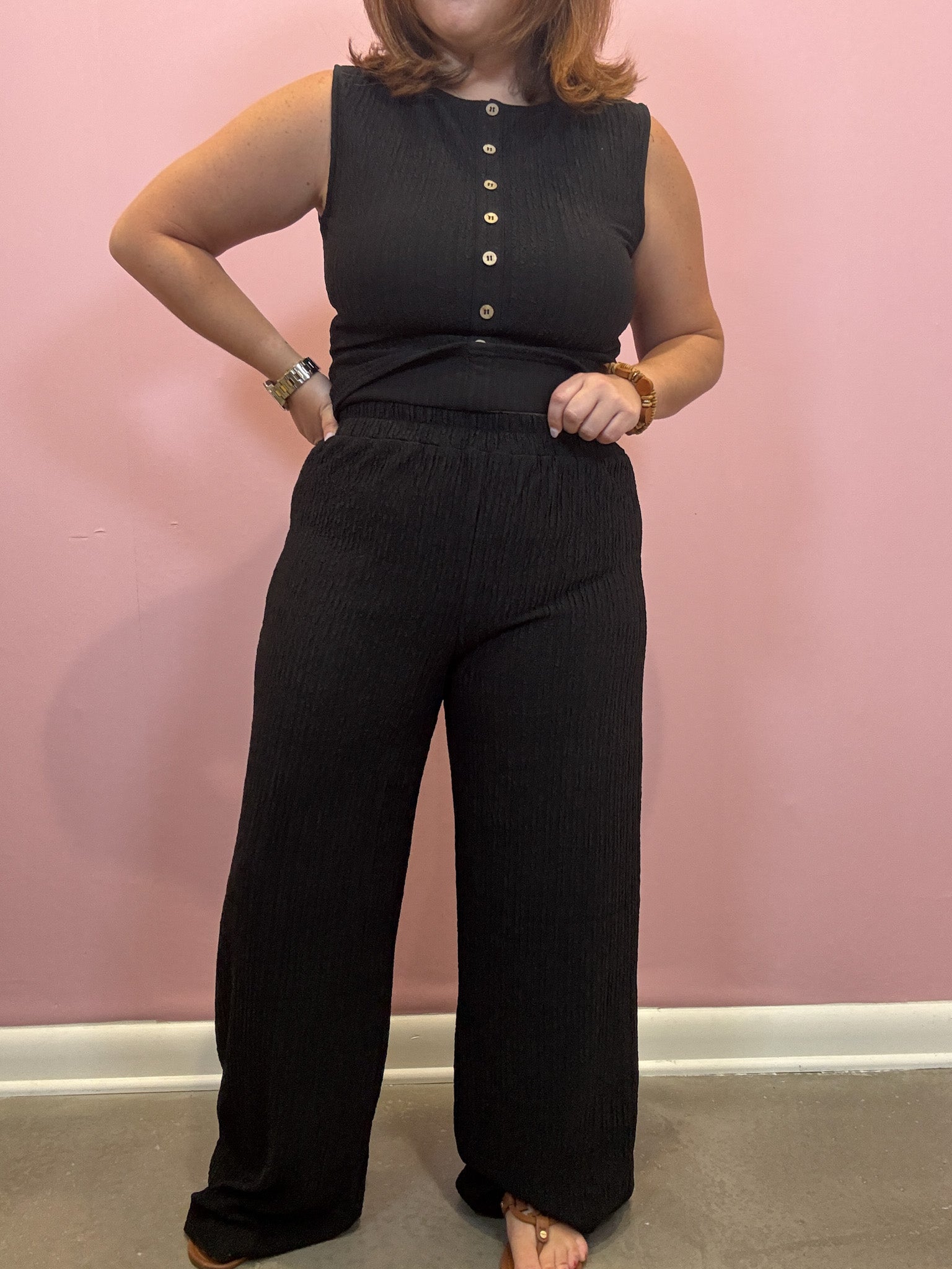 A woman modeling a black ribbed sleeveless top and matching wide-leg pants set against a pink wall.