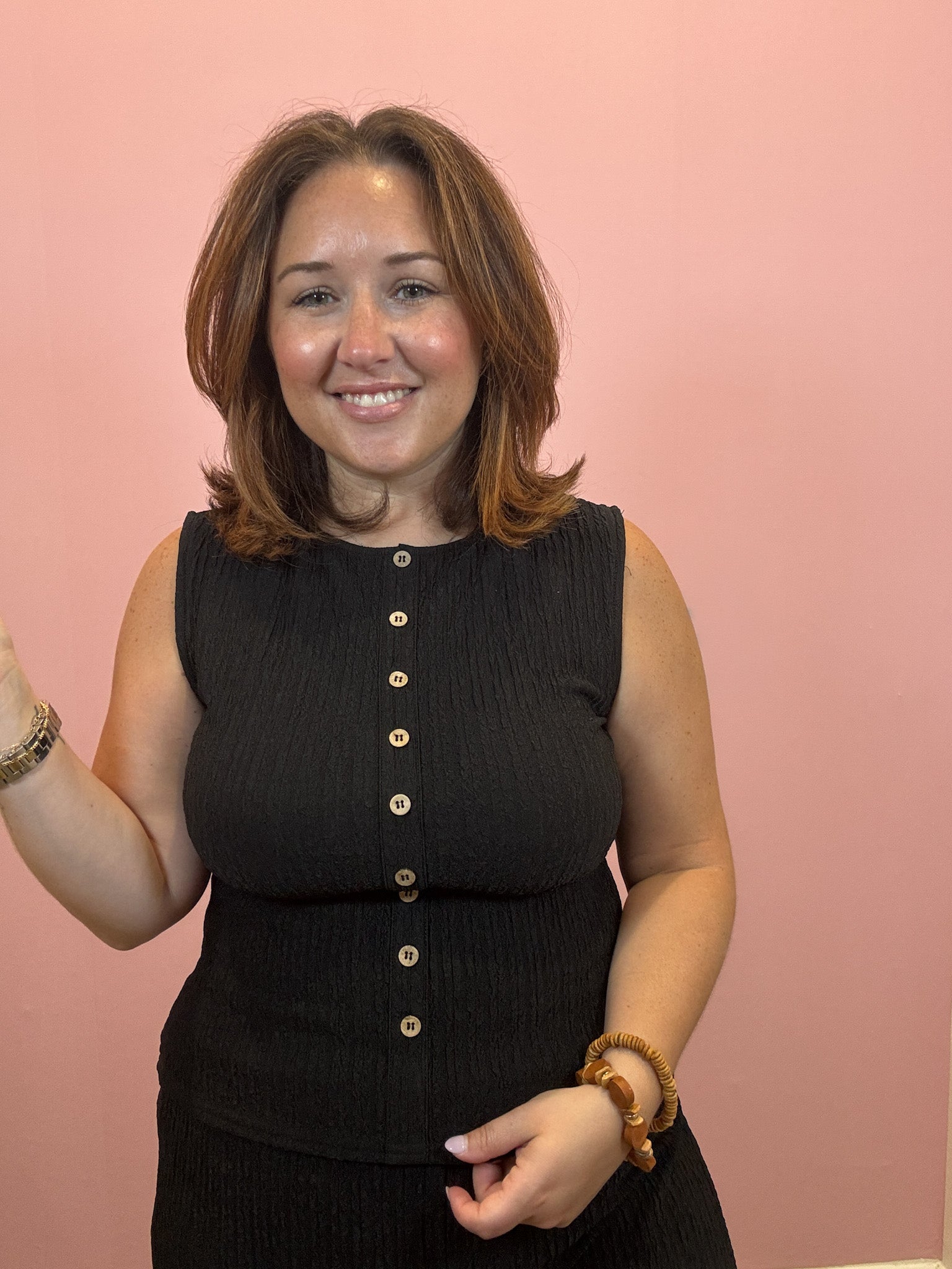 A woman with shoulder-length brown hair smiling in front of a solid pink wall, wearing a sleeveless black ribbed top with gold buttons down the front.
