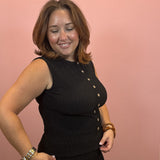 A woman modeling a sleeveless black ribbed top with gold buttons down the front, posing against a solid pink background.