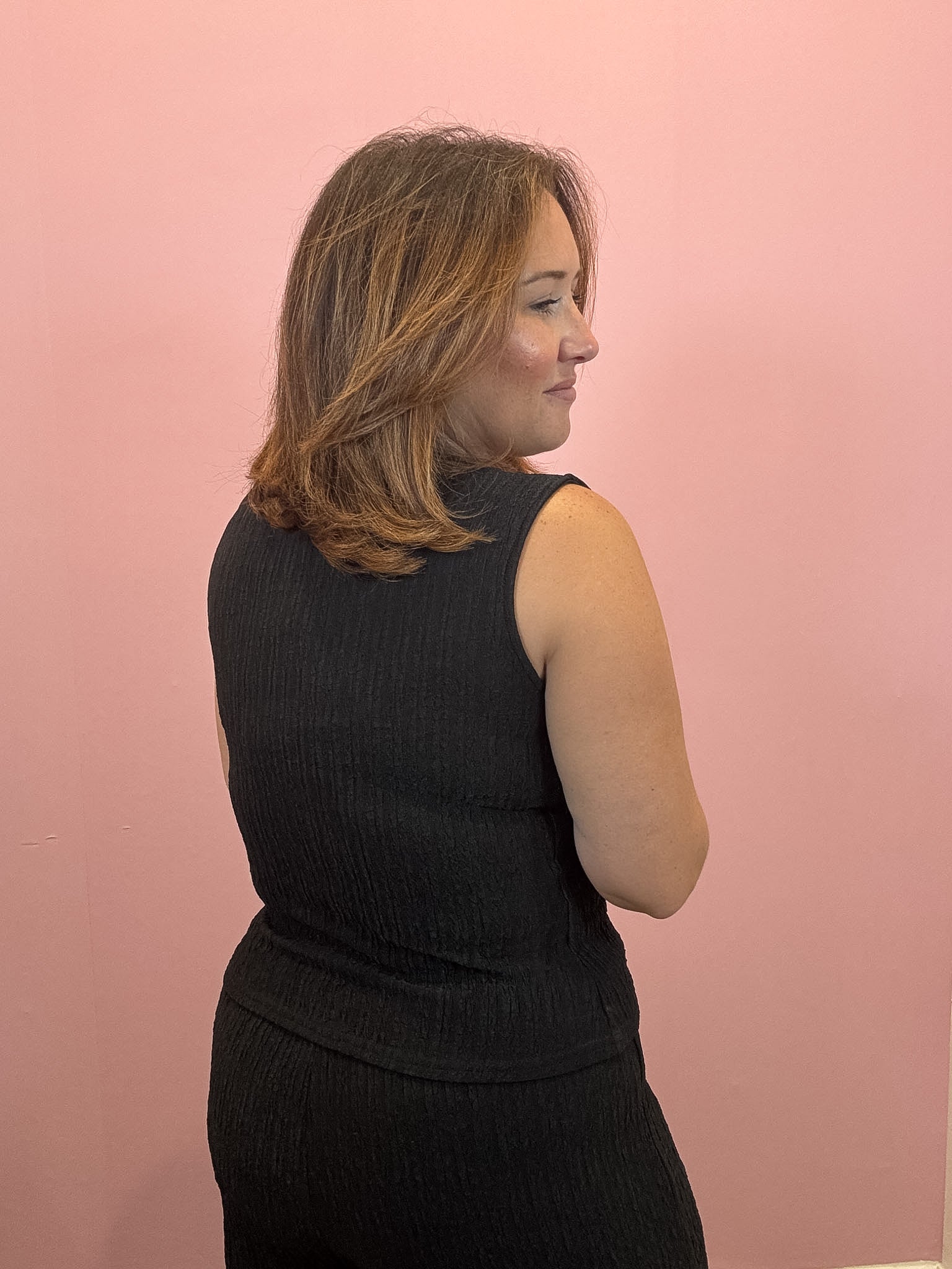 A woman with shoulder-length brown hair is shown from the back and side, wearing a sleeveless black ribbed top against a solid pink wall.