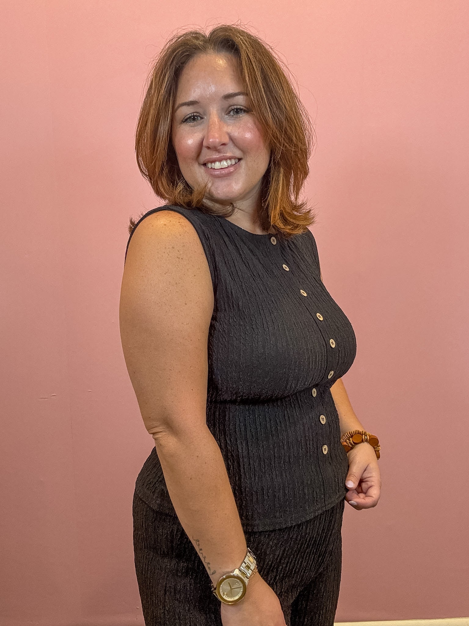 A woman with shoulder-length brown hair smiling and posing in a sleeveless dark gray ribbed top with gold buttons, set against a solid dusty pink wall.