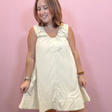 A woman modeling a sleeveless cream-colored dress with a gathered neckline and ruffled hem, standing against a solid pink wall.