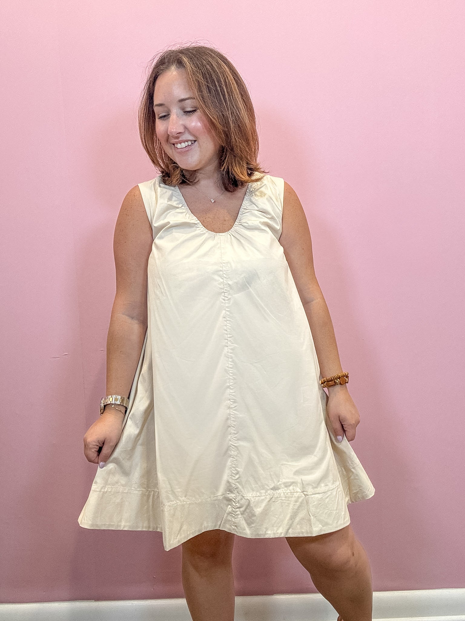 A woman modeling a sleeveless cream-colored dress with a gathered neckline and ruffled hem, standing against a solid pink wall.