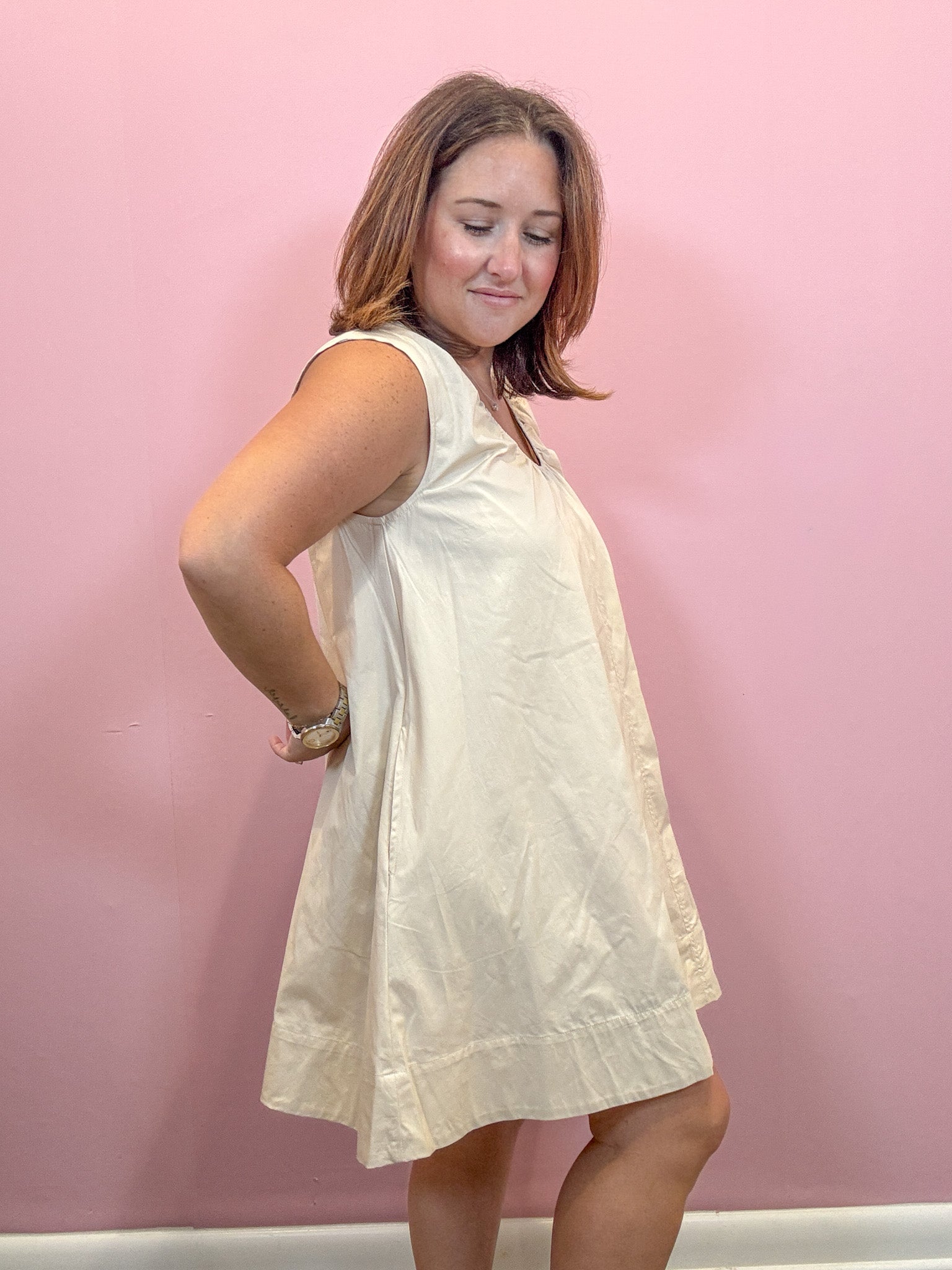 A woman modeling a sleeveless, A-line mini dress in a warm off-white or cream color, standing against a solid pink wall.