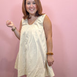 A woman with shoulder-length brown hair smiling and wearing a sleeveless cream-colored dress against a solid pink wall.
