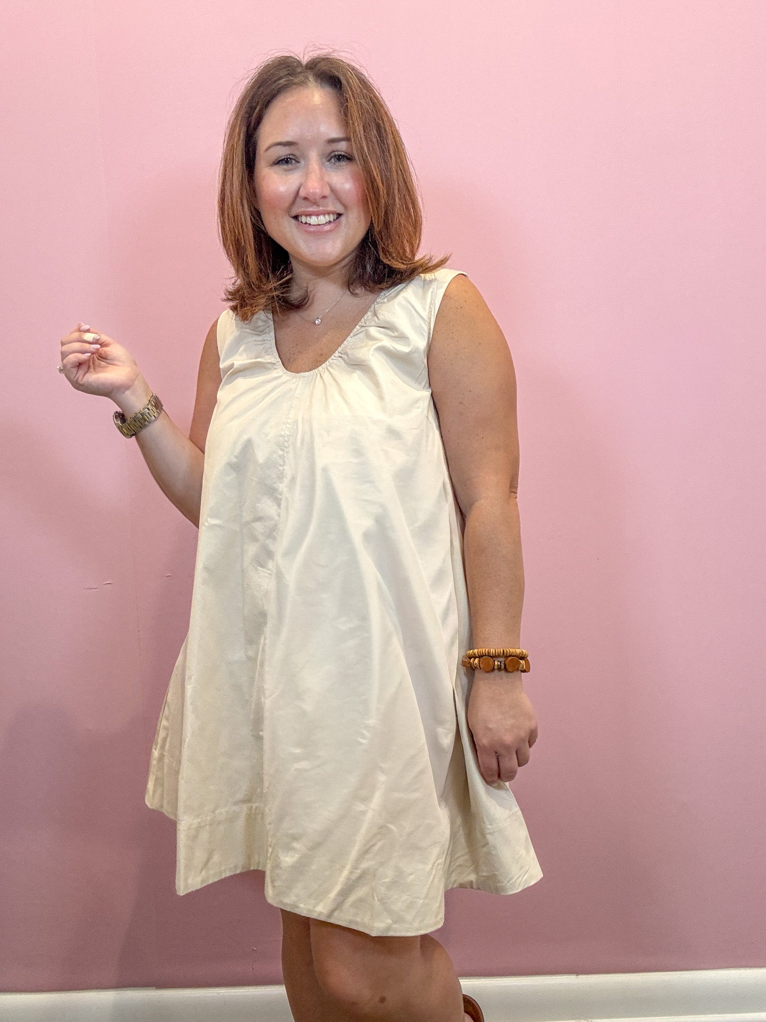 A woman with shoulder-length brown hair smiling and wearing a sleeveless cream-colored dress against a solid pink wall.