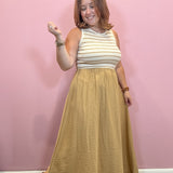 A woman modeling a sleeveless maxi dress with a cream and tan striped top and a textured mustard yellow skirt, standing against a pink wall.