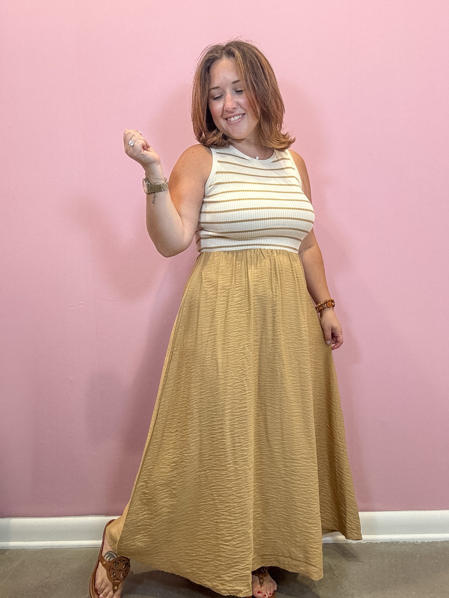 A woman modeling a sleeveless maxi dress with a cream and tan striped top and a textured mustard yellow skirt, standing against a pink wall.