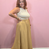 A woman modeling a sleeveless top and long skirt set against a solid pink wall. The top is cream with thin mustard yellow horizontal stripes, and the matching skirt is a textured mustard yellow.