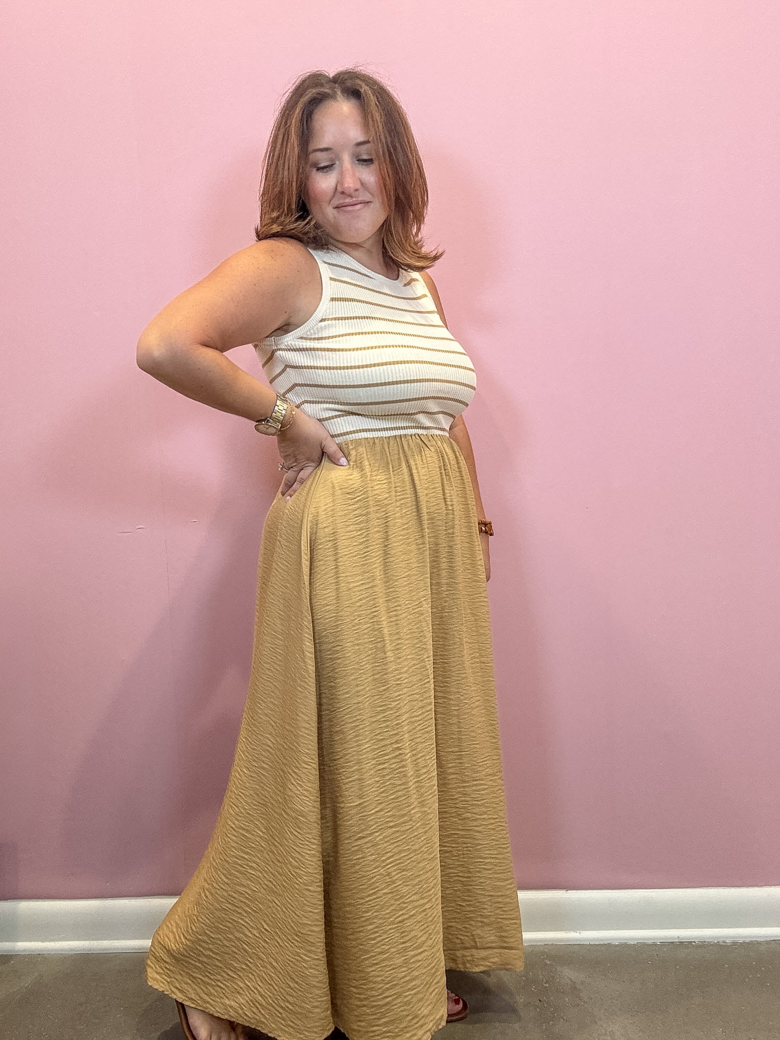A woman modeling a sleeveless top and long skirt set against a solid pink wall. The top is cream with thin mustard yellow horizontal stripes, and the matching skirt is a textured mustard yellow.