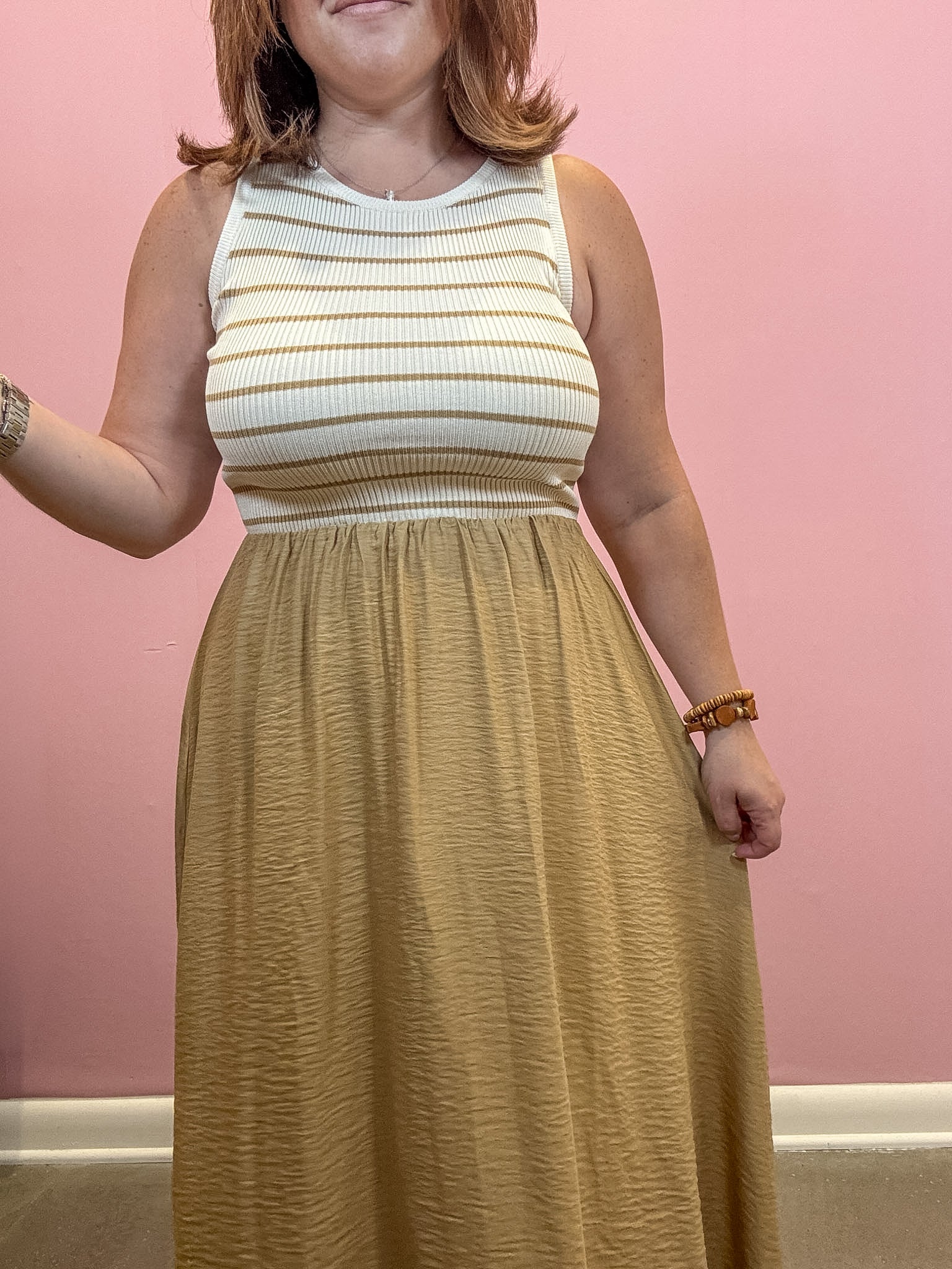 A woman wearing a sleeveless ribbed knit maxi dress with a cream and tan striped bodice and a flowing mustard-gold skirt, standing against a pink wall.