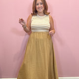 A woman modeling a sleeveless maxi dress with a cream and tan striped bodice and a textured mustard-gold skirt, standing against a solid pink wall.