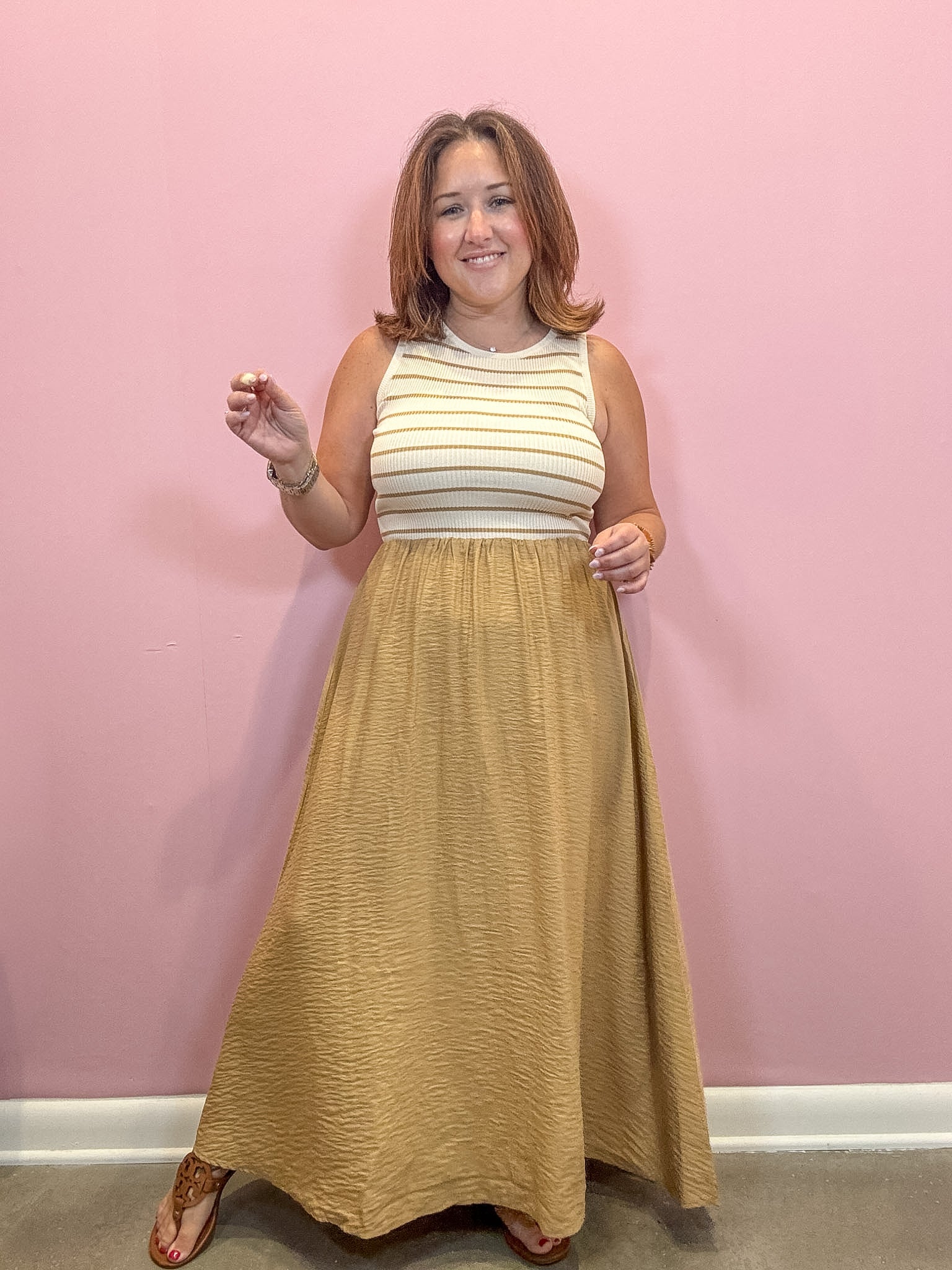 A woman modeling a sleeveless maxi dress with a cream and tan striped bodice and a textured mustard-gold skirt, standing against a solid pink wall.