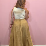A woman stands with her back to the camera wearing a sleeveless white top with thin mustard yellow horizontal stripes and a long, flowing mustard yellow skirt, set against a solid pink wall.
