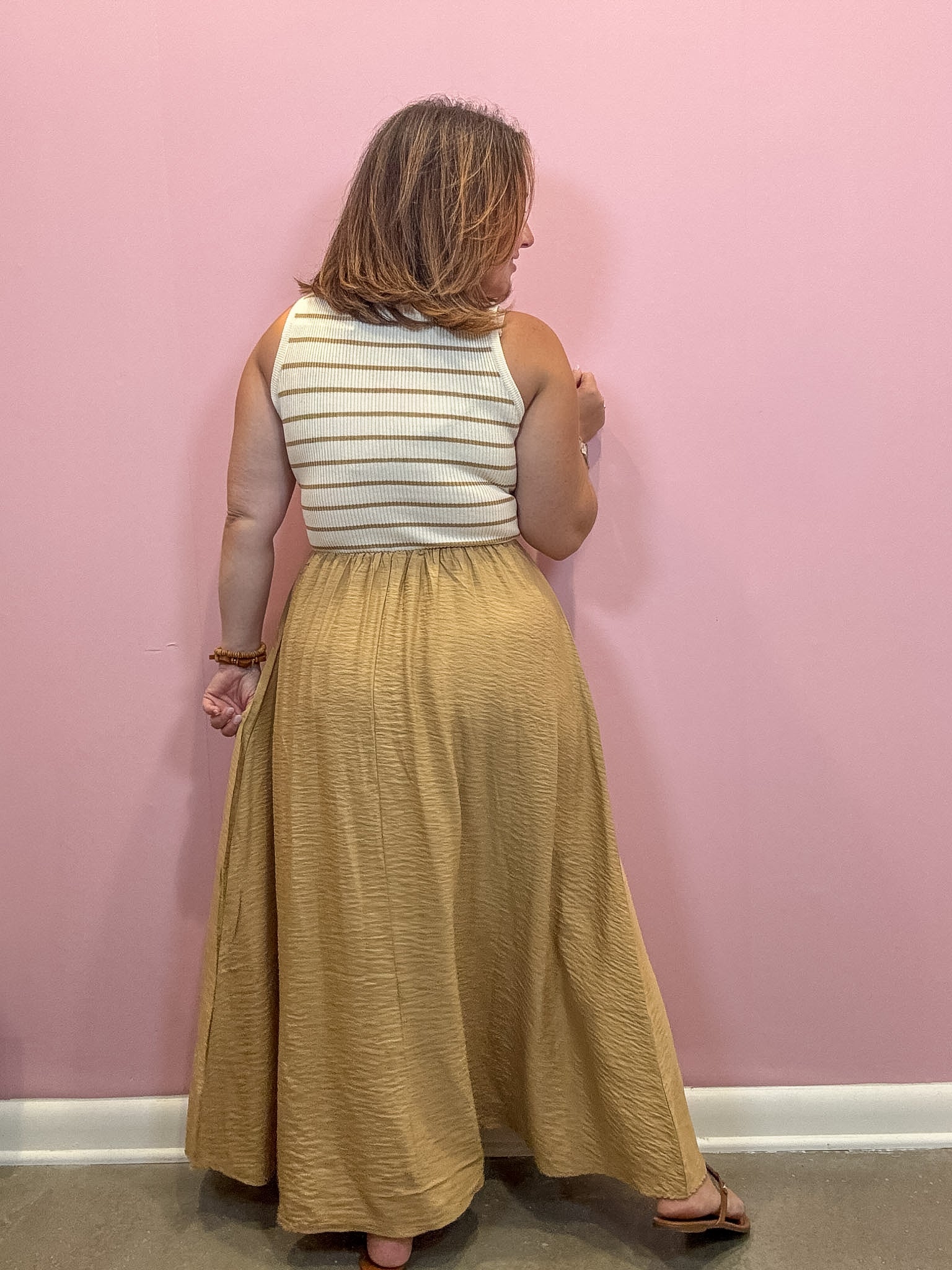 A woman stands with her back to the camera wearing a sleeveless white top with thin mustard yellow horizontal stripes and a long, flowing mustard yellow skirt, set against a solid pink wall.