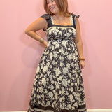 A woman modeling a black and cream floral maxi dress with ruffled cap sleeves, posing against a solid pink wall.