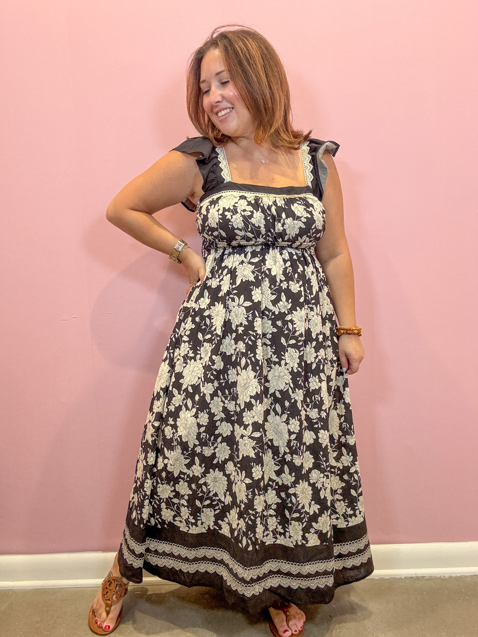 A woman modeling a black and cream floral maxi dress with ruffled cap sleeves, posing against a solid pink wall.