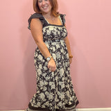 A woman modeling a black and white floral maxi dress with ruffled cap sleeves, standing against a solid pink wall.