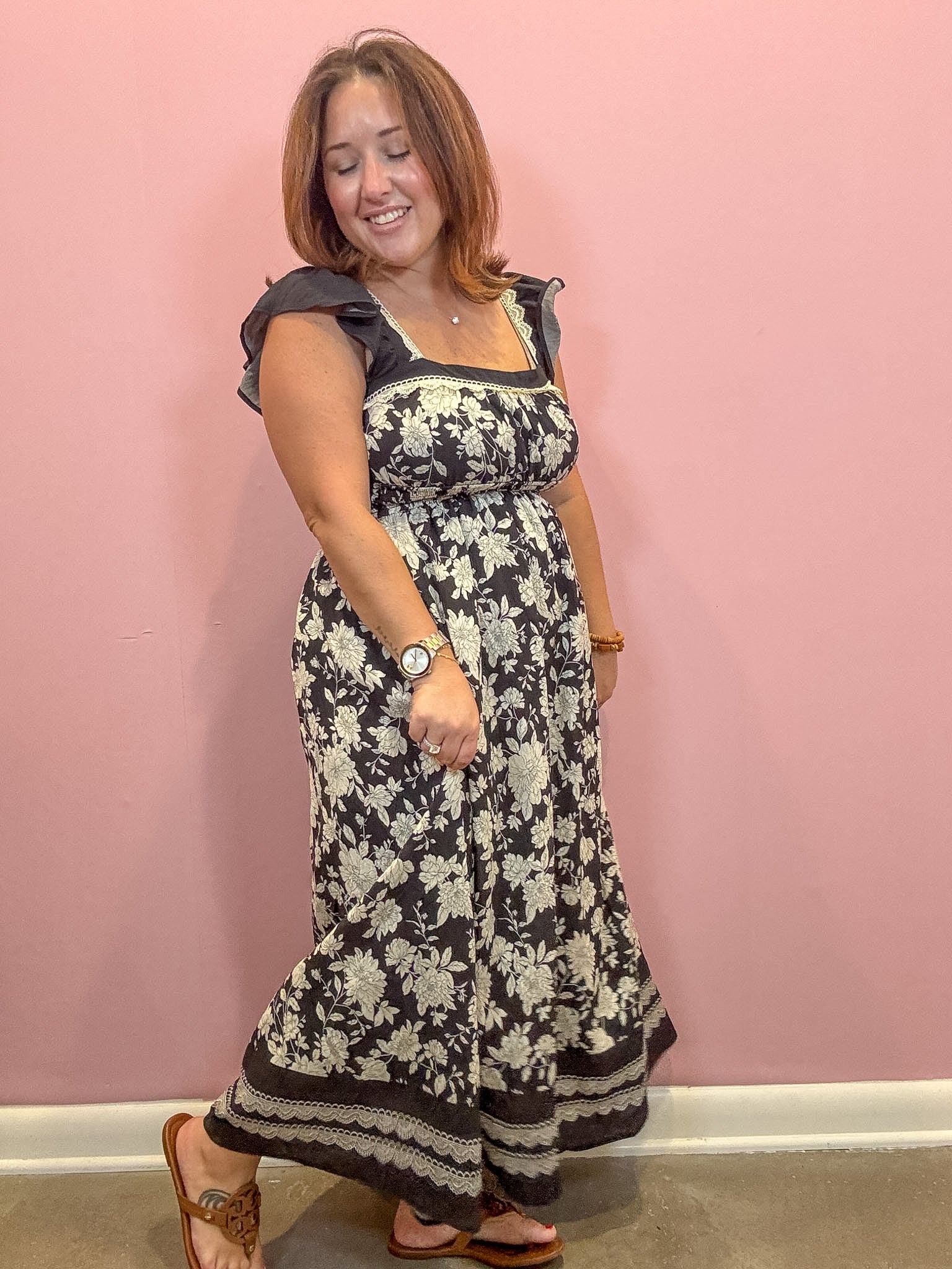 A woman modeling a black and white floral maxi dress with ruffled cap sleeves, standing against a solid pink wall.