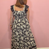 A woman viewed from behind wearing a sleeveless black maxi dress with a cream floral print, ruffled shoulder straps, and a smocked waist, standing against a solid pink wall.