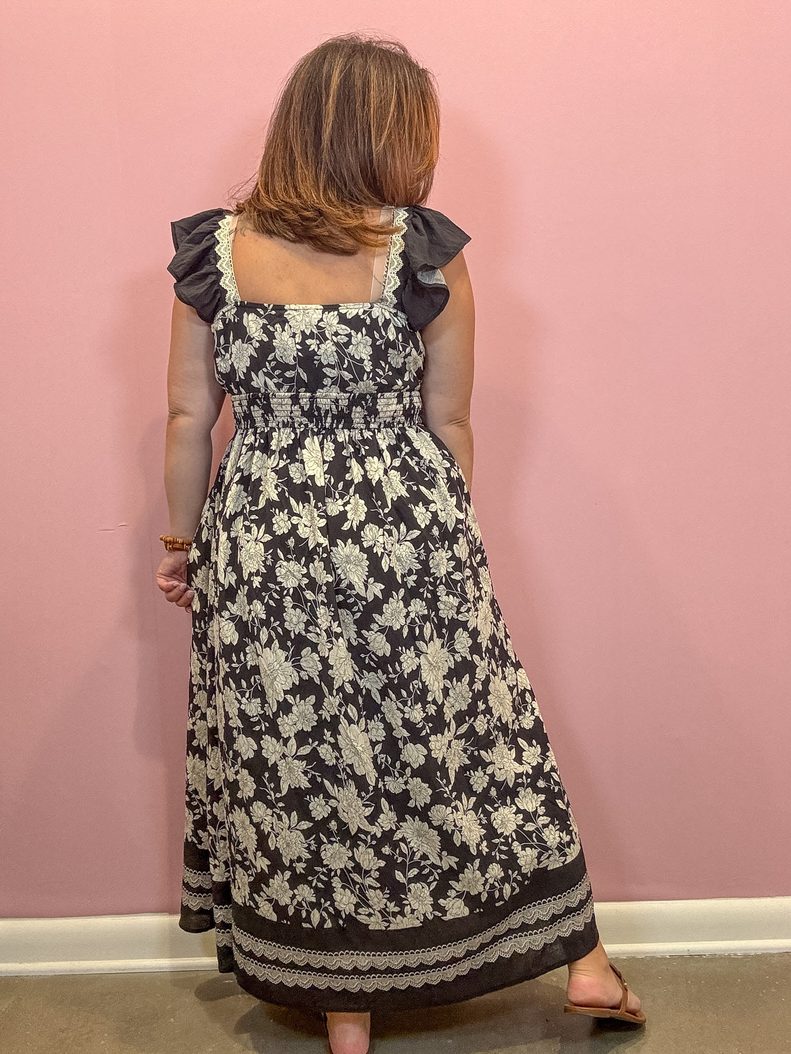 A woman viewed from behind wearing a sleeveless black maxi dress with a cream floral print, ruffled shoulder straps, and a smocked waist, standing against a solid pink wall.