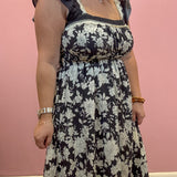 A woman wearing a black midi dress with a white floral pattern, ruffled cap sleeves, and a smocked bodice, standing against a solid pink wall.