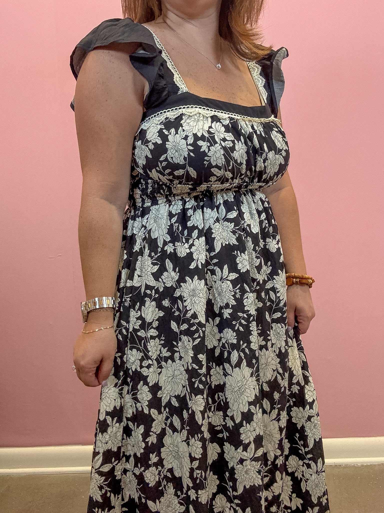 A woman wearing a black midi dress with a white floral pattern, ruffled cap sleeves, and a smocked bodice, standing against a solid pink wall.