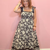 A woman modeling a sleeveless black and cream floral maxi dress with ruffled shoulder straps and a scalloped hem, standing against a solid pink wall.