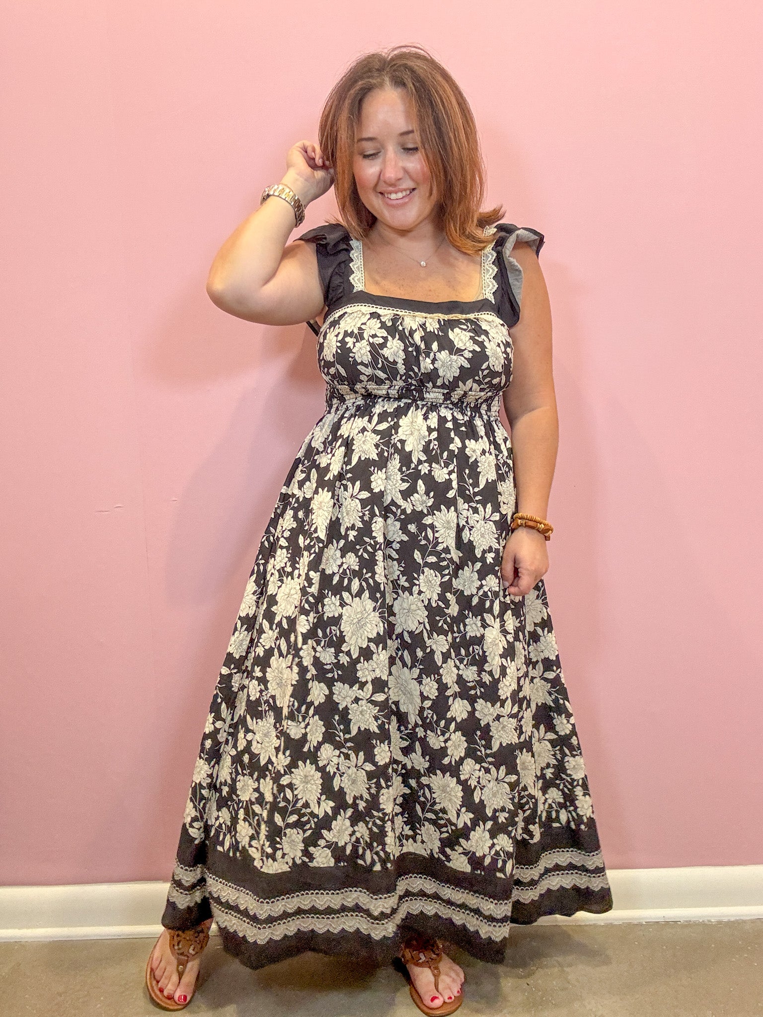 A woman modeling a sleeveless black and cream floral maxi dress with ruffled shoulder straps and a scalloped hem, standing against a solid pink wall.