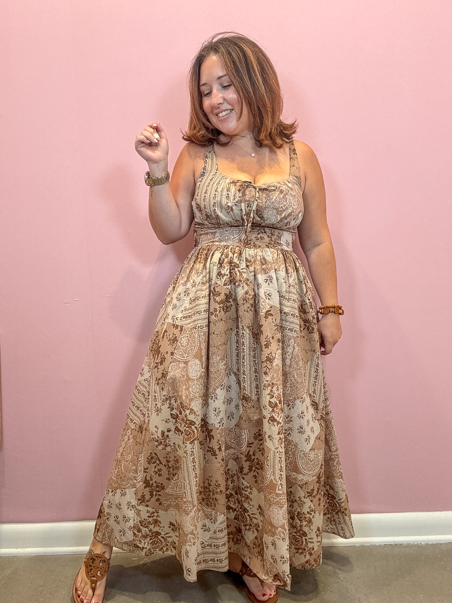 A woman modeling a sleeveless, floor-length maxi dress with a beige and brown paisley pattern, standing against a solid pink wall.