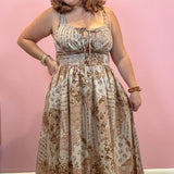 A woman wearing a sleeveless maxi dress with a beige and brown paisley print, standing against a solid pink wall.