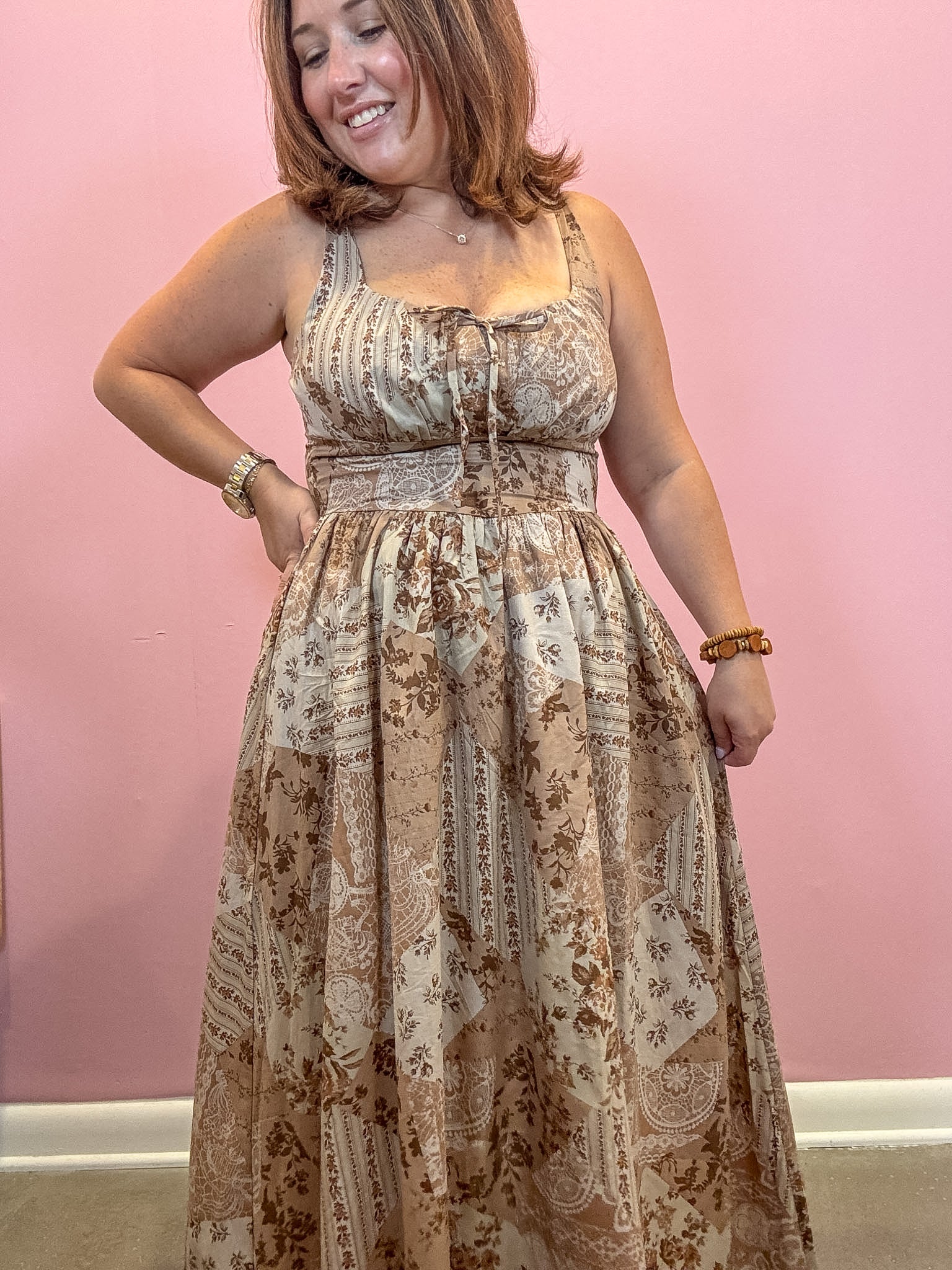 A woman wearing a sleeveless maxi dress with a beige and brown paisley print, standing against a solid pink wall.