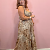 A woman modeling a sleeveless, floor-length maxi dress with a beige and brown paisley pattern, standing against a solid pink wall.