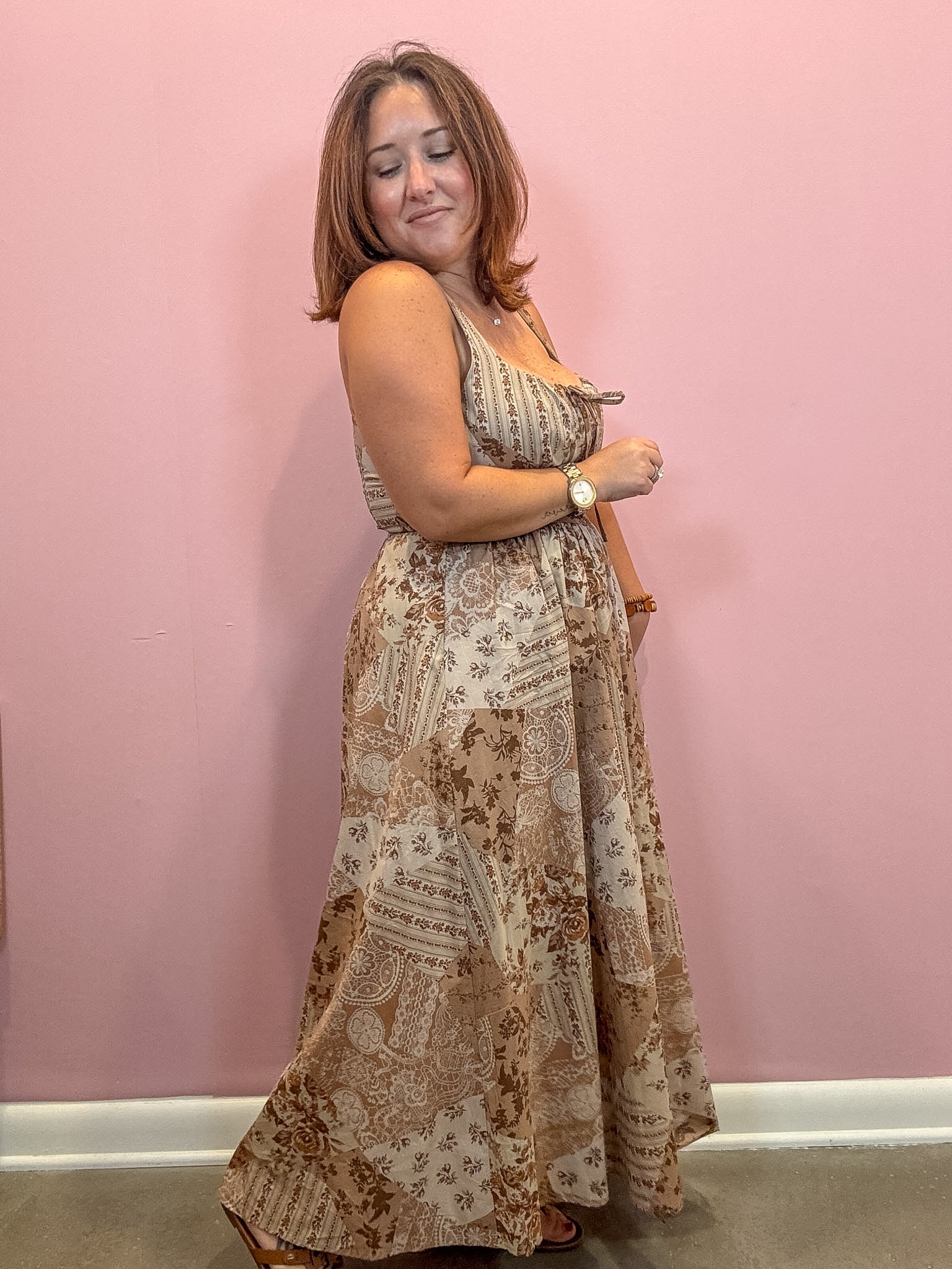 A woman modeling a sleeveless, floor-length maxi dress with a beige and brown paisley pattern, standing against a solid pink wall.