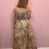 A woman viewed from behind wearing a sleeveless maxi dress with a beige and brown patchwork-style toile print, standing against a solid pink wall.