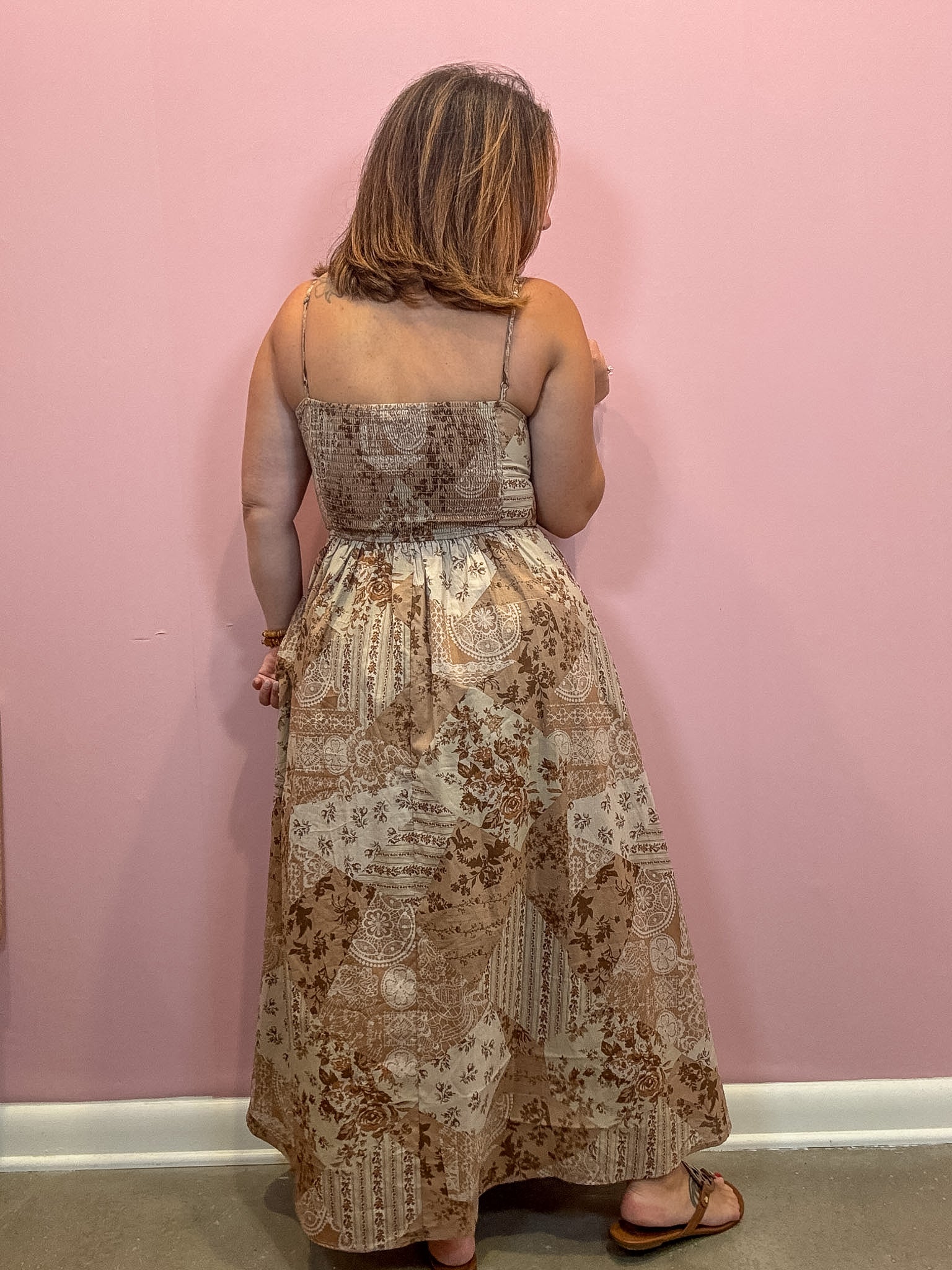 A woman viewed from behind wearing a sleeveless maxi dress with a beige and brown patchwork-style toile print, standing against a solid pink wall.