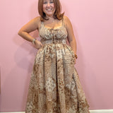 A woman modeling a sleeveless, floor-length maxi dress with a beige and brown vintage script and floral pattern, standing against a solid pink wall.