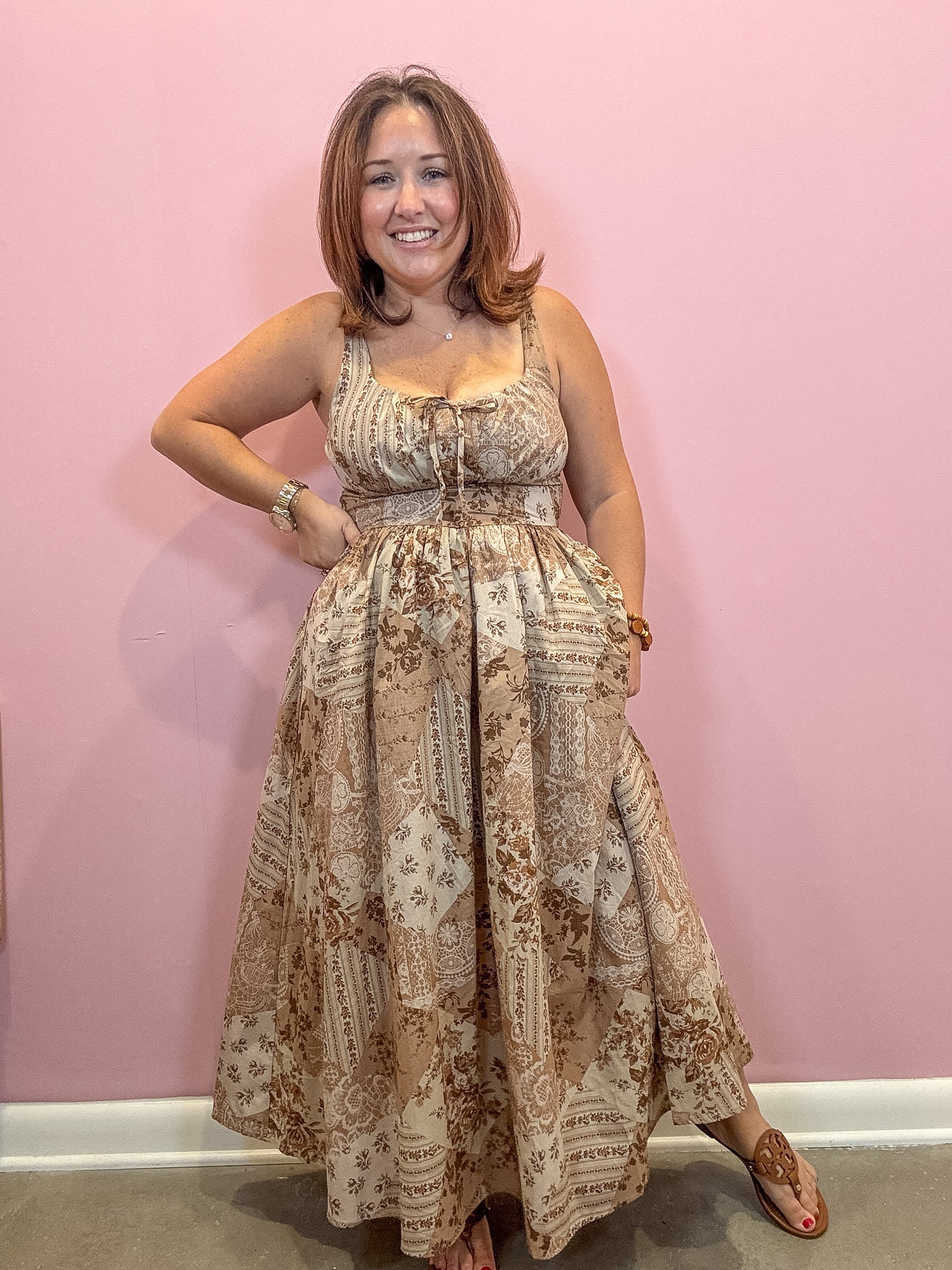 A woman modeling a sleeveless, floor-length maxi dress with a beige and brown vintage script and floral pattern, standing against a solid pink wall.