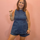 A woman modeling a sleeveless blue denim romper with matching denim shorts against a solid pink wall.