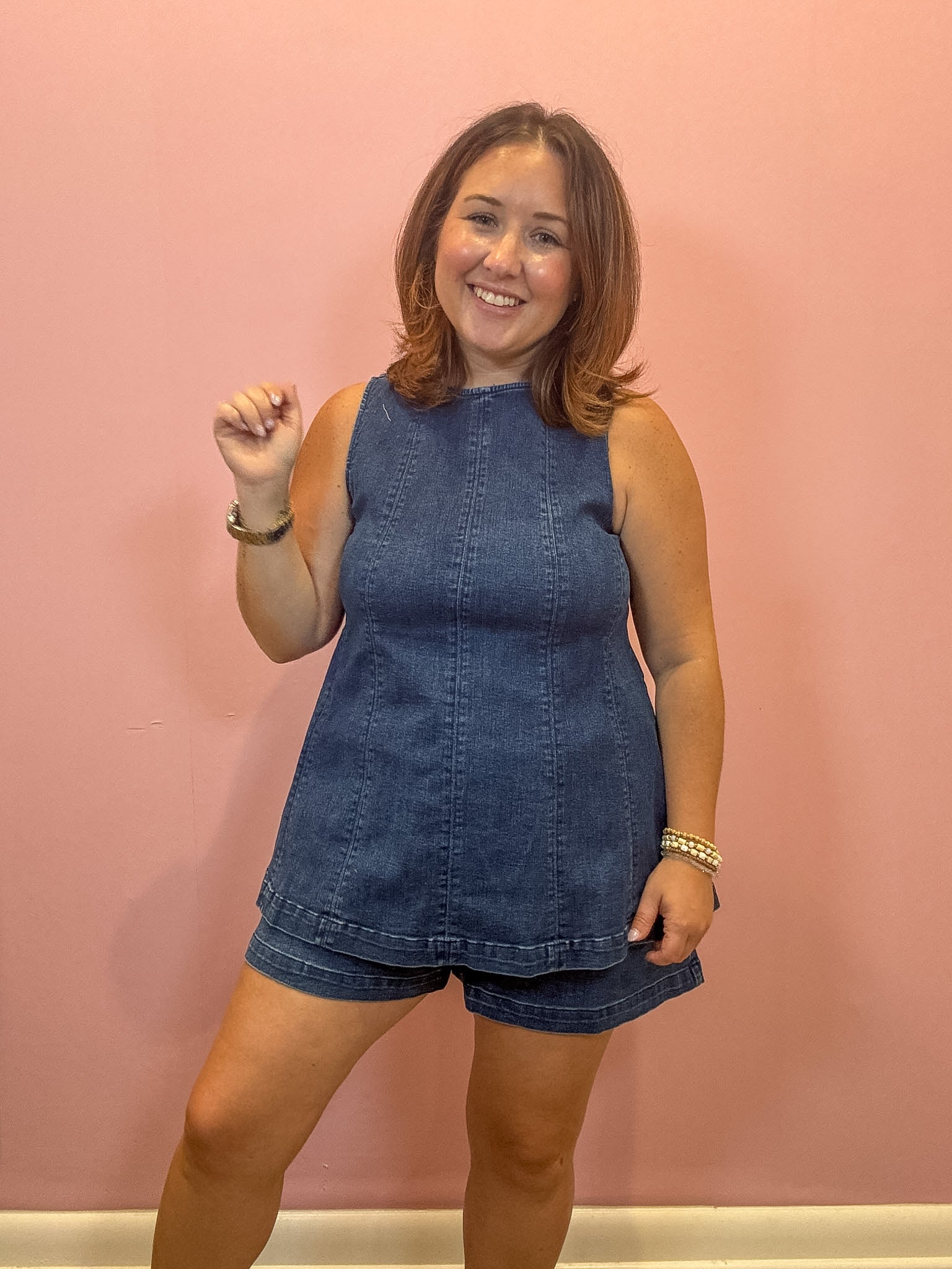 A woman modeling a sleeveless blue denim romper with matching denim shorts against a solid pink wall.