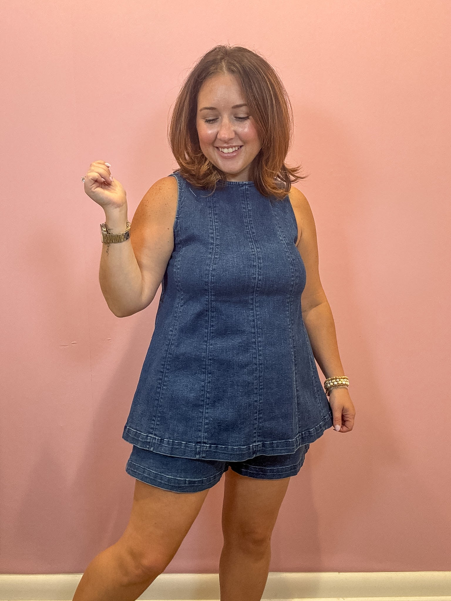 A woman modeling a sleeveless blue denim top and matching denim shorts against a solid pink wall.