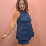 A woman modeling a sleeveless blue denim tunic top and matching denim shorts against a solid pink wall.