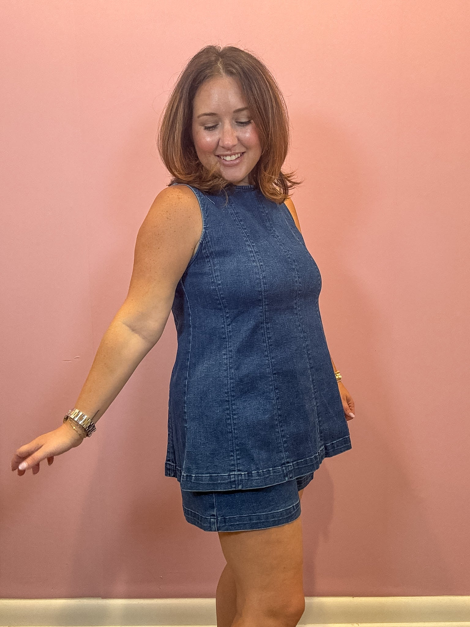 A woman modeling a sleeveless blue denim tunic top and matching denim shorts against a solid pink wall.