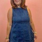 A woman modeling a sleeveless dark blue denim mini dress with a tiered skirt, standing against a solid pink wall.