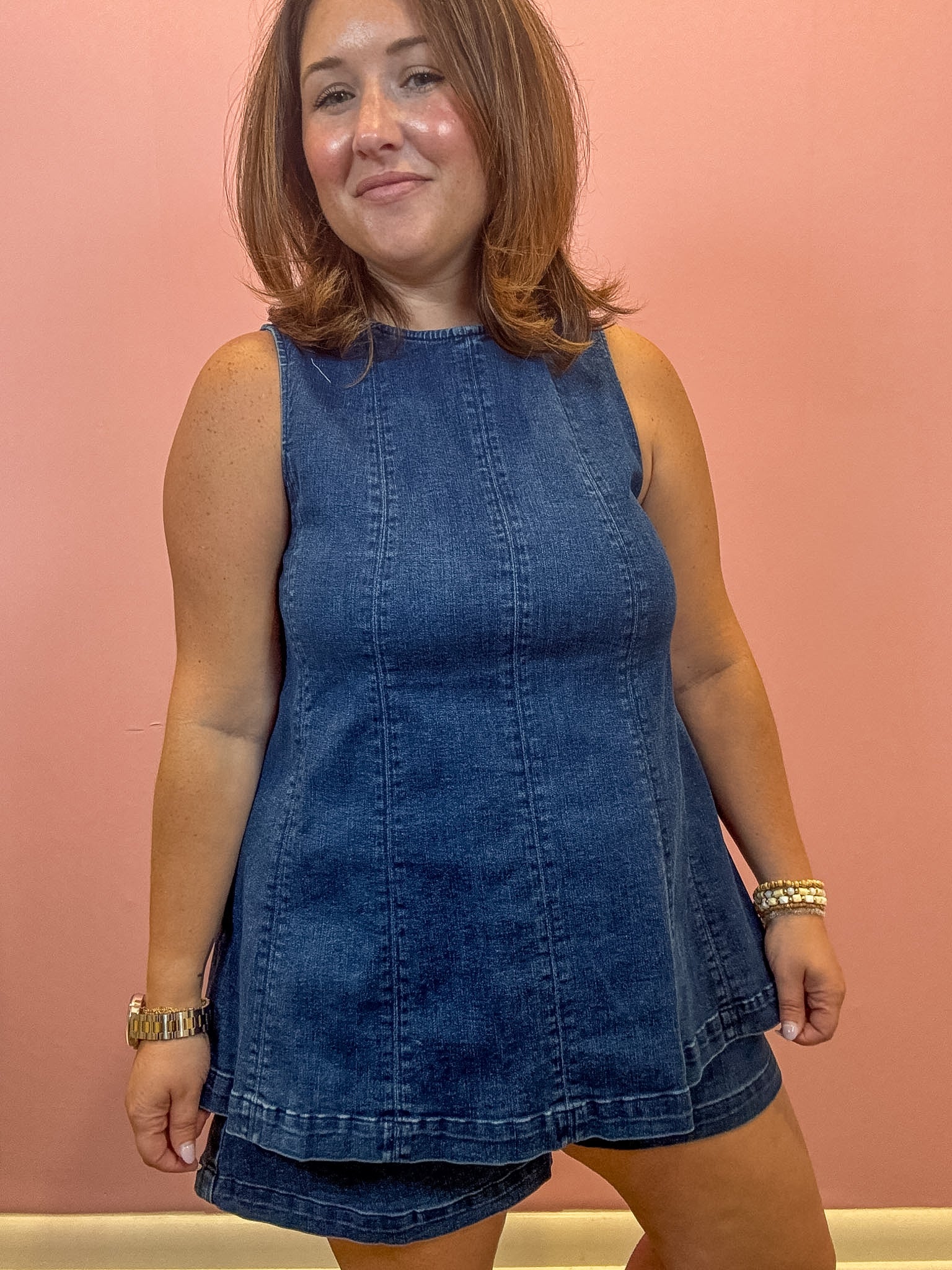 A woman modeling a sleeveless dark blue denim mini dress with a tiered skirt, standing against a solid pink wall.