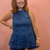 A woman modeling a sleeveless blue denim romper with matching denim shorts, standing against a solid pink wall.